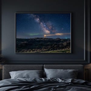 Könnte beinhalten: Ein rechteckiger gerahmter Kunstdruck mit einer nächtlichen Landschaft, einer hellen Milchstraße, Sternen und einem leuchtenden Leuchtturmstrahl über einem dunklen Horizont, zentriert über einem grauen Polsterbett in einem dunklen Raum.