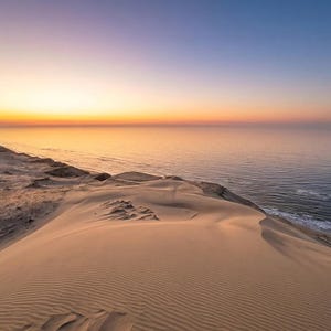 Könnte beinhalten: Landschaftsfotografie einer Sanddüne bei Sonnenuntergang. Die Düne befindet sich im Vordergrund, mit dem Meer und einem farbenfrohen Himmel im Hintergrund. Der Himmel wechselt von Blau zu Orange und Gelb.