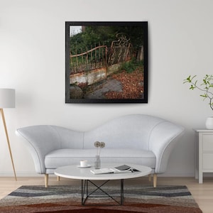 May include: A square framed wall art print featuring a weathered, rusted metal fence set against a backdrop of dark green foliage. The ground in front of the fence is covered in fallen brown autumn leaves, with a small section of gray stone paving.