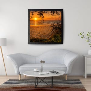Könnte beinhalten: Ein quadratisches gerahmtes Wandbild, das einen Sonnenuntergang über einem Strand in Orange- und Gelbtönen zeigt, mit Silhouetten von Baumzweigen im Vordergrund und ruhigen Meereswellen am sandigen Ufer bei Dämmerung.