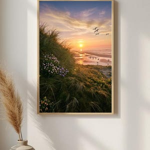 Könnte beinhalten: Ein gerahmter Kunstdruck mit einer Küstenlandschaft bei Sonnenuntergang. Das Bild zeigt grasbewachsene Sanddünen mit kleinen violetten Blumen im Vordergrund, einen Sandstrand und eine leuchtend orangefarbene Sonne über dem Ozean.