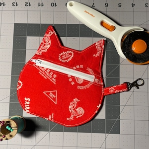 Könnte beinhalten: Eine rote Stofftasche in Katzenform mit Reißverschluss und einem weißen Hahn-Design. Die Tasche trägt den Text "Sriracha" und "Fong Foods, Inc.". Die Tasche liegt auf einer grauen Schneidematte mit einem Rollschneider und einer Garnrolle.