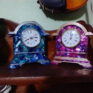 May include: Two decorative mantel clocks with white clock faces and Roman numerals. One clock has a blue and green mosaic design, the other has a pink and purple mosaic design. Both clocks have silver and gold accents.