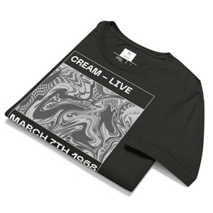 May include: Black t-shirt featuring a graphic design with the text "CREAM - LIVE" and "MARCH 7TH 1968" in white. The graphic is a swirling, abstract pattern in shades of gray and white. The shirt is folded.