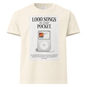 Pode incluir: Camiseta creme com um gráfico de um iPod e o texto "1.000 SONGS in your POCKET." O gráfico inclui o texto "Um reprodutor de música MP3 inovador que armazena até 100 músicas de qualidade em um design ultraportátil de 184 gramas que cabe no seu bolso."
