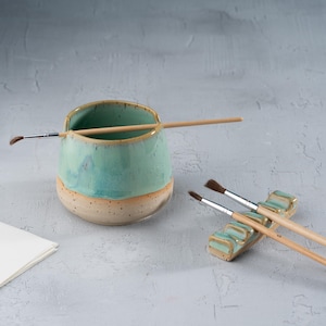 May include: A ceramic brush holder with a turquoise glaze and a speckled beige base. A paintbrush rests in the holder, and two paintbrushes are on a matching brush rest. The art supplies are on a light gray surface.