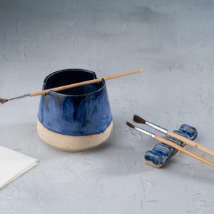 May include: A handmade ceramic art studio set. The set includes a deep blue and cream-coloured ceramic brush holder with a paintbrush resting on top, and a small blue brush rest with two paintbrushes. A stack of white paper is in the corner.