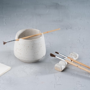 May include: A speckled white ceramic brush holder with a wide opening, holding a paintbrush. Two additional paintbrushes rest on a small, white ceramic brush rest. A few sheets of white paper are visible on the left.