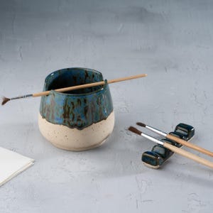 May include: A ceramic brush holder with a two-tone design. The top half is glazed in shades of blue and green, while the bottom is a creamy white. A paintbrush rests in the holder, and a small brush rest holds two additional paintbrushes.