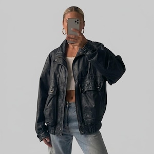 May include: A black leather bomber jacket featuring a front zipper closure, a high collar, and two large flap pockets on the chest. The jacket has long sleeves with ribbed cuffs and a ribbed hem, worn over a white tank top and light blue denim jeans.