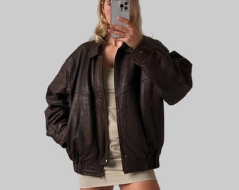 May include: A dark brown leather jacket with a classic bomber style. The jacket features a collared neckline, button closures, and ribbed cuffs and hem. The leather appears worn, giving it a vintage aesthetic. The jacket is a medium size.