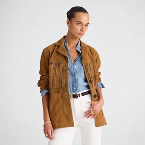 May include: A tan suede jacket with fringing on the chest and sleeves. The jacket features a collared neckline, button fastenings, and two front pockets. The model is wearing a denim shirt and white trousers.
