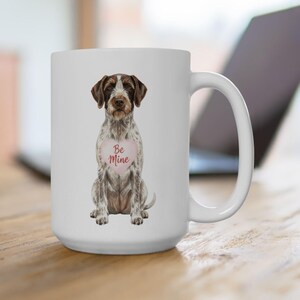 May include: White ceramic mug featuring a seated dog illustration with brown and white fur. The dog has a pink heart-shaped tag that reads "Be Mine". The mug has a large handle and is likely used for beverages.