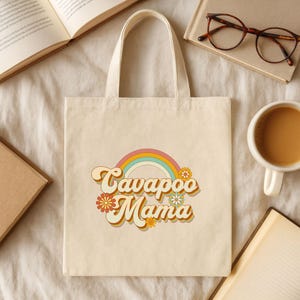 May include: Cream-colored tote bag with the text "Cavapoo Mama" in a retro font, arched over a rainbow and flowers. The bag has a simple design with a rectangular shape and two handles.