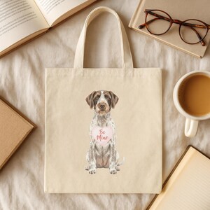 May include: Cream-colored tote bag featuring a watercolor illustration of a dog with brown and white fur. The dog has a pink heart-shaped sign that says "Be Mine". The bag has a simple design with two handles.