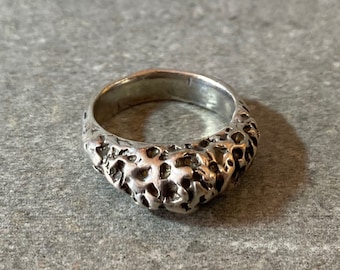 Puede incluir: Un anillo de plata con un diseño texturizado y orgánico. El anillo está hecho de metal y tiene un acabado liso y pulido.