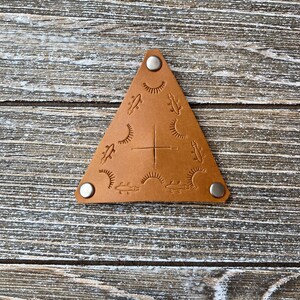 May include: A triangular leather keychain with a brown leather background and silver metal accents. The keychain features a design of stylized lizards and sunbursts.
