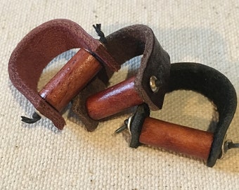 May include: Three leather and wood finger rings. The rings are brown, black, and burgundy. Each ring has a wooden dowel that is attached to the leather band.