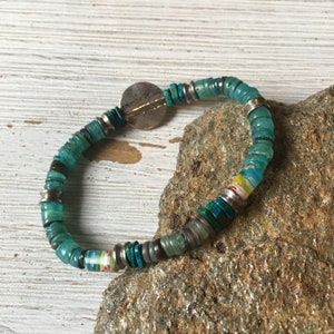 May include: A turquoise and silver beaded bracelet with a round silver charm. The bracelet is made of various shades of turquoise and silver beads.