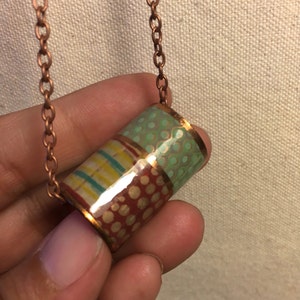 Pode incluir: Colar de cobre feito à mão com um pendente cilíndrico. O pendente é decorado com padrões coloridos, incluindo listras, bolinhas e um design pontilhado em vermelho, verde e amarelo. A corrente também é de cobre.