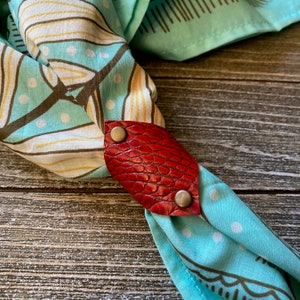 Könnte beinhalten: Ein roter Leder-Bandana-Halter mit zwei silbernen Druckknöpfen. Der Halter ist an einem türkisfarbenen Bandana mit weißen Punkten befestigt.