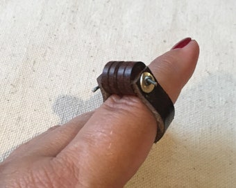May include: A brown leather ring with metal accents. The ring is adjustable and has a unique design.