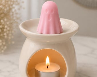 May include: A cream-colored ceramic wax warmer with a wide top dish and a hollow base containing a lit tea light candle. A small, light pink wax melt shaped like a ghost with two oval eyes and an open mouth sits on the top dish of the warmer.