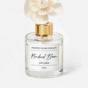 May include: A clear glass diffuser bottle with a gold cap and a large, cream-colored flower on top. The label reads "Kindred Glow Candles" and "Kindred Bloom Diffuser 100ml".