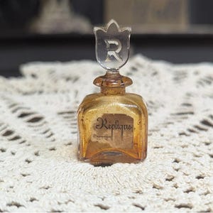 May include: A small, vintage perfume bottle with a clear, tulip-shaped stopper featuring the letter "R". The bottle is square with a golden hue and a label that reads "Replique". The bottle is displayed on a white, lace-like surface.