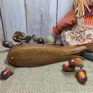 Könnte beinhalten: Ein hölzernes fischförmiges Objekt mit geschnitzten Schuppendetails, das auf einer Jutefläche liegt. Eicheln und Tannenzapfen umgeben den Fisch, mit einer Vogelscheuchenpuppe im Hintergrund. Es ist ein dekoratives Objekt.