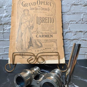 May include: An antique opera libretto for "Carmen" with a vintage brass stand, next to a pair of silver binoculars and a silver telescope. The libretto is printed on aged paper and features ornate text and illustrations.