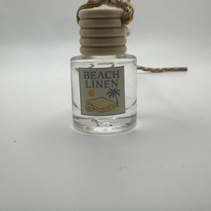 Könnte beinhalten: Eine klare Glasflasche mit einem Holzdeckel und einer dekorativen Schnur. Die Flasche hat ein Etikett mit der Aufschrift "BEACH LINEN" mit einer Grafik eines Handtuchs und einer Palme. Die Flasche ist als Lufterfrischer konzipiert.