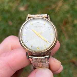 May include: Vintage Accutron watch with a gold-tone case and a silver dial. The watch features gold hands and hour markers, with diamond accents at the 3, 6, 9, and 12 o'clock positions. The watch band is a gold-tone, expandable style.