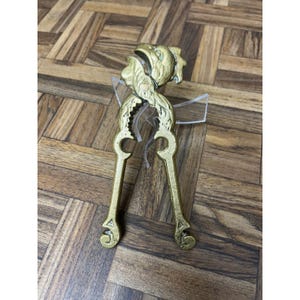 May include: A gold-colored nutcracker with a decorative design. The nutcracker has a detailed head and handles with a curved design. The nutcracker is made of metal and is designed to crack nuts.