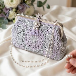 May include: A crocheted clutch in shades of lavender and silver, adorned with sequins and a pearl-beaded chain. The clasp is silver, and a decorative floral embellishment with pearls and a tassel adds elegance. A bouquet of flowers is in the background.