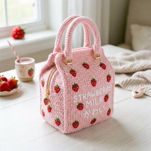 Puede incluir: Un bolso de mano rosa tejido a ganchillo con fresas rojas bordadas. El bolso tiene una cremallera y asas doradas, con las palabras "STRAWBERRY MILK" y caracteres japoneses bordados en la parte delantera. El bolso mide unos 20 cm de alto.