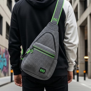 Op de afbeelding: Grijze gehaakte sling bag met neon groene rits- en banddetails. De tas heeft een klein, vierkant logo en een patroonband. De tas wordt over de schouder gedragen.