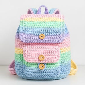 Op de afbeelding: Een handgemaakte gehaakte rugzak in regenboog pastelkleuren. De tas heeft een klepsluiting met een houten knoop, twee voorzakken met houten knopen en verstelbare bandjes. Een zacht, textuurrijk en kleurrijk accessoire.
