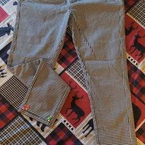 May include: Black and white houndstooth patterned pants with a drawstring waist. The pants have colorful accents at the pockets and cuffs. The pants are displayed on a patterned surface with bears, deer, and plaid.
