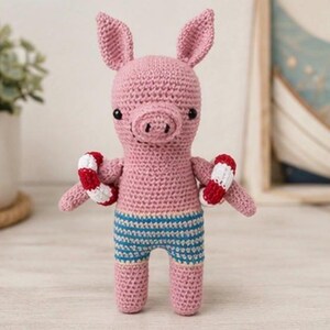 May include: A pink crocheted pig toy featuring two pointed ears, a snout, and black bead eyes. The pig is wearing blue and white striped shorts and has a red and white striped life preserver ring around each of its two arms.