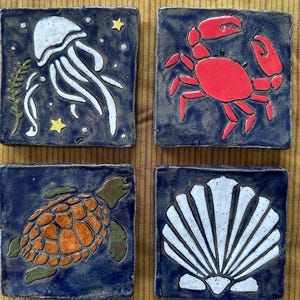 May include: Set of four square ceramic tiles with sea creature designs. Includes a white jellyfish, a red crab, an orange and green sea turtle, and a white seashell, all on a dark blue background.