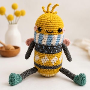 May include: A crochet bee plush toy featuring a yellow head with black eyes, pink cheeks, and antennae. The body has a blue and white striped section, a dark gray middle with white diamond patterns, and dark gray limbs with teal feet.
