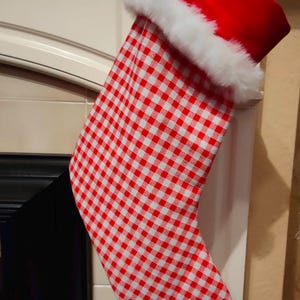 Può includere: Una calza di Natale con un motivo a quadri rossi e bianchi. La parte superiore è rossa con un bordo in finta pelliccia bianca. La calza è appesa a un caminetto bianco. Un anello a quadri viene utilizzato per appenderla.