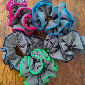 Op de afbeelding: Een verzameling grijze scrunchies met kleurrijke randen. De scrunchies hebben een gerimpeld ontwerp en zijn afgewerkt met felroze, blauwe en groene randen. Ze liggen op een houten oppervlak.