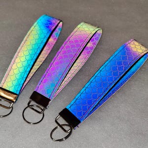 May include: Three wristlet keychains featuring a reflective, iridescent scale pattern. Each strap is secured with a metal clamp and a circular split ring. The colours shift between blue, green, purple, and yellow hues across the textured surfaces.