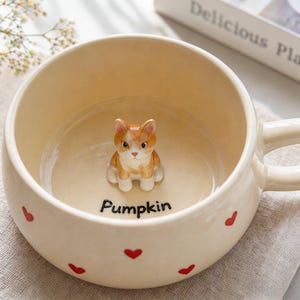 Puede incluir: Taza de cerámica color crema con una pequeña figura de gato naranja y blanco en el interior. La taza tiene un asa y está decorada con formas de corazón rojas. La palabra "Pumpkin" está escrita en negro en el interior de la taza.