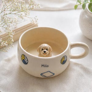 May include: Cream-colored ceramic mug with a sculpted puppy inside. The mug has a handle and is decorated with blue and yellow designs and the name "Milo". The puppy has a light brown coat and is sticking its tongue out.