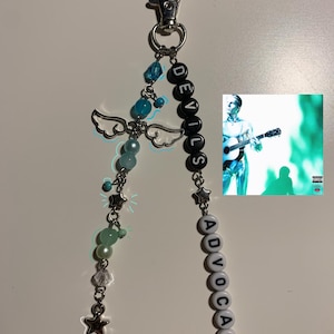 May include: A keychain featuring a silver lobster clasp, two strands of beads, and a small square image of a person playing guitar. One strand has blue and green beads with silver angel wings and star charms. The other has black and white letter beads spelling DEVILS ADVOCATE.