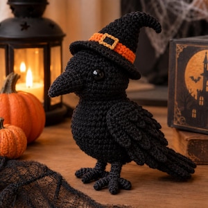 Könnte beinhalten: Eine schwarze, gehäkelte Rabenfigur mit einem spitzen schwarzen Hexenhut, der mit einem orangefarbenen Band und einer gelben Schnalle verziert ist. Der Vogel hat einen strukturierten Garnkörper, einen spitzen Schnabel und glänzende Augen.