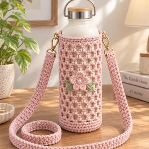 May include: A white water bottle with a silver and wood lid, encased in a light pink crocheted sleeve featuring a lattice pattern, a central pink flower applique with a pearl center, green leaf accents, and a matching pink crocheted shoulder strap.